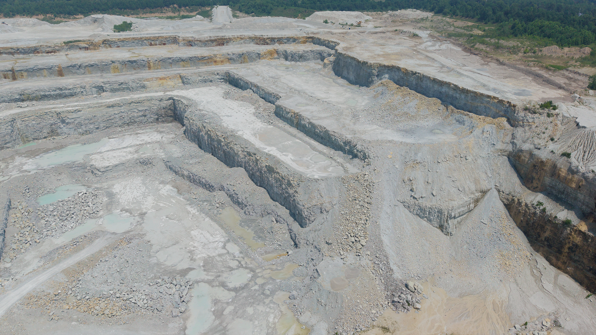 Aerial view of an active quarry pit with stepped benches, high vertical highwalls, and exposed rock strata captured for pit-progression documentation