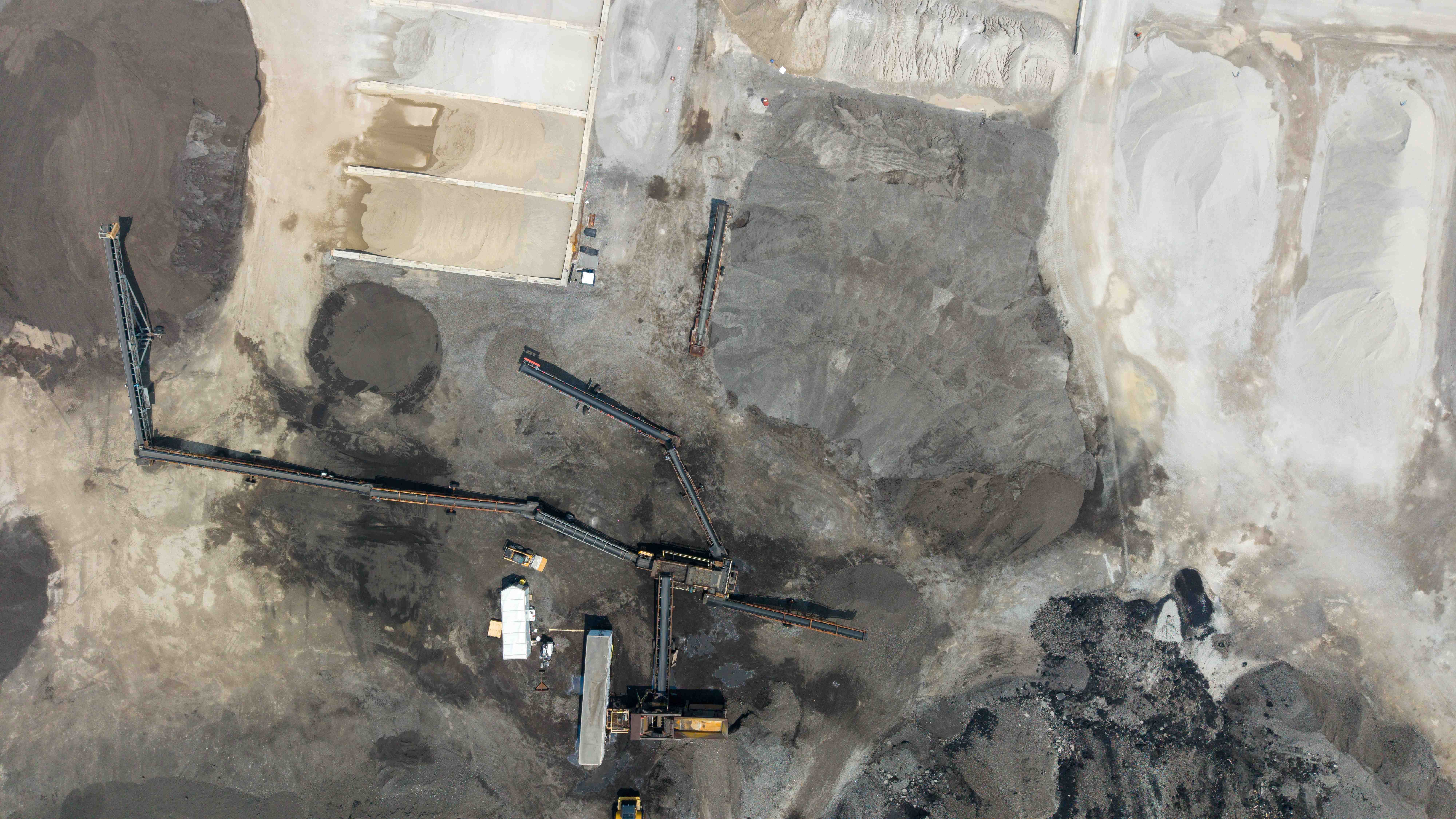 Top-down aerial of an active aggregate processing yard showing radial stacker and conveyor lines, graded sand and material stockpiles, a wheel loader, and plant equipment