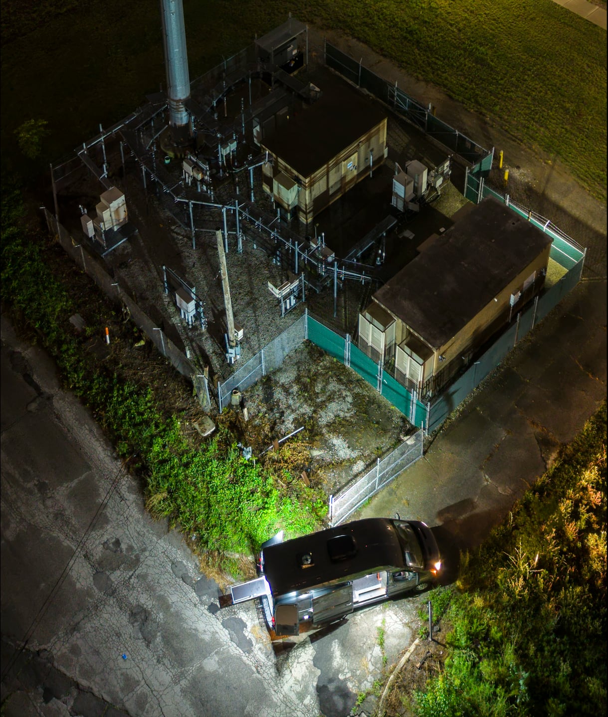 Overhead night aerial of a fenced telecom compound showing the monopole tower, equipment shelters, and cabinets, with the MIR command van staged at the entrance under floodlight