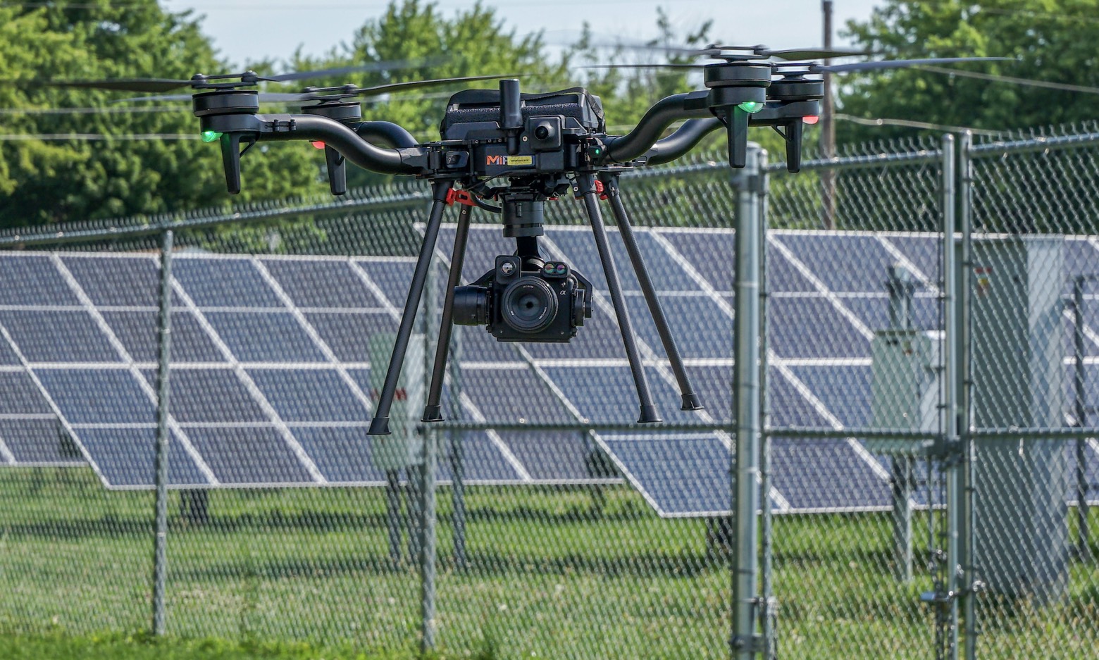 MIR drone with a gimbal-mounted thermal and RGB payload hovering in front of a ground-mounted solar array behind a security fence