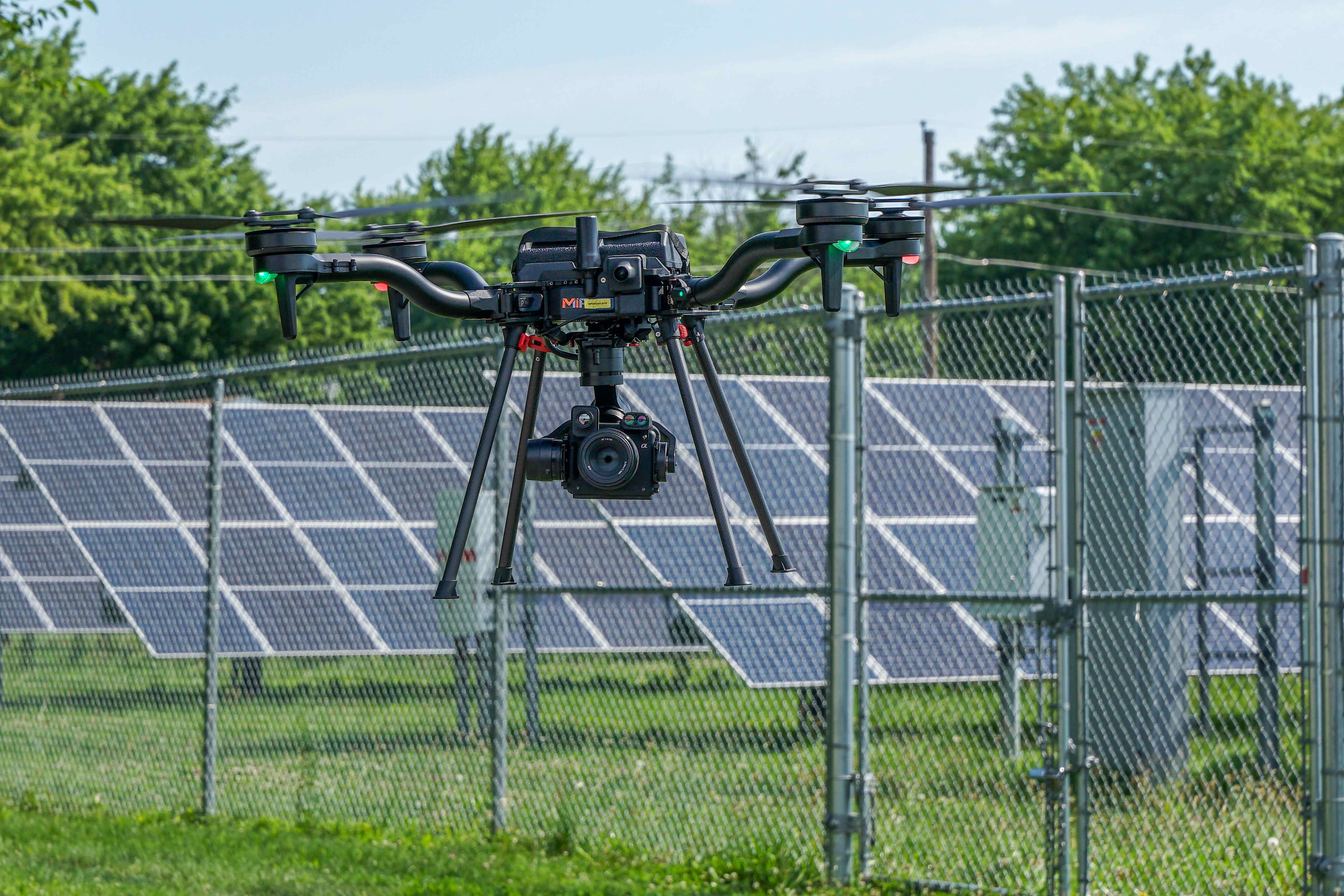 MIR Freefly Astro aircraft with RGB and thermal gimbal payload in flight in front of a fenced ground-mounted solar array