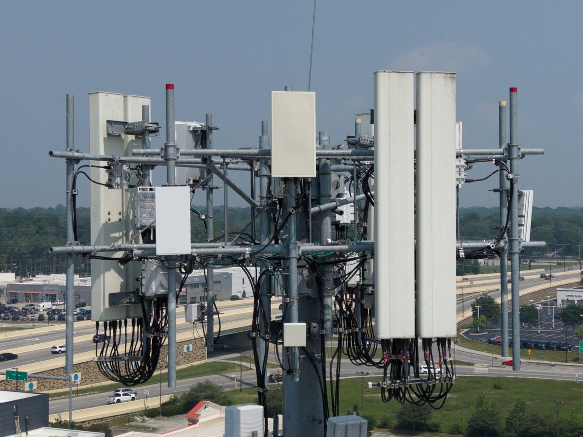 Visible-light aerial photo of a monopole cell tower loaded with panel antennas, remote radio units, and coax cabling above a highway interchange