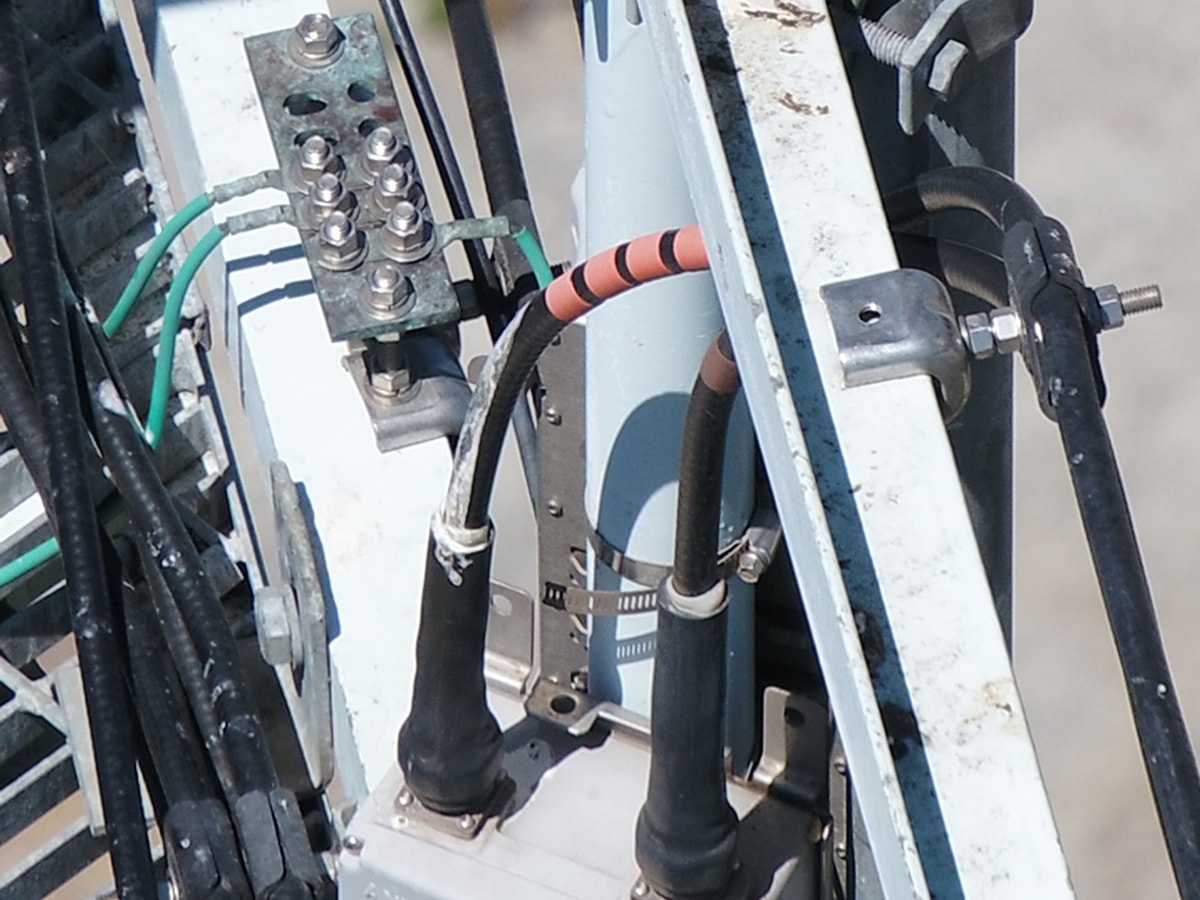 High-resolution close-up of a cell tower antenna base showing the grounding bus bar, bolts, ground wires, and weatherproofed coax jumper connectors