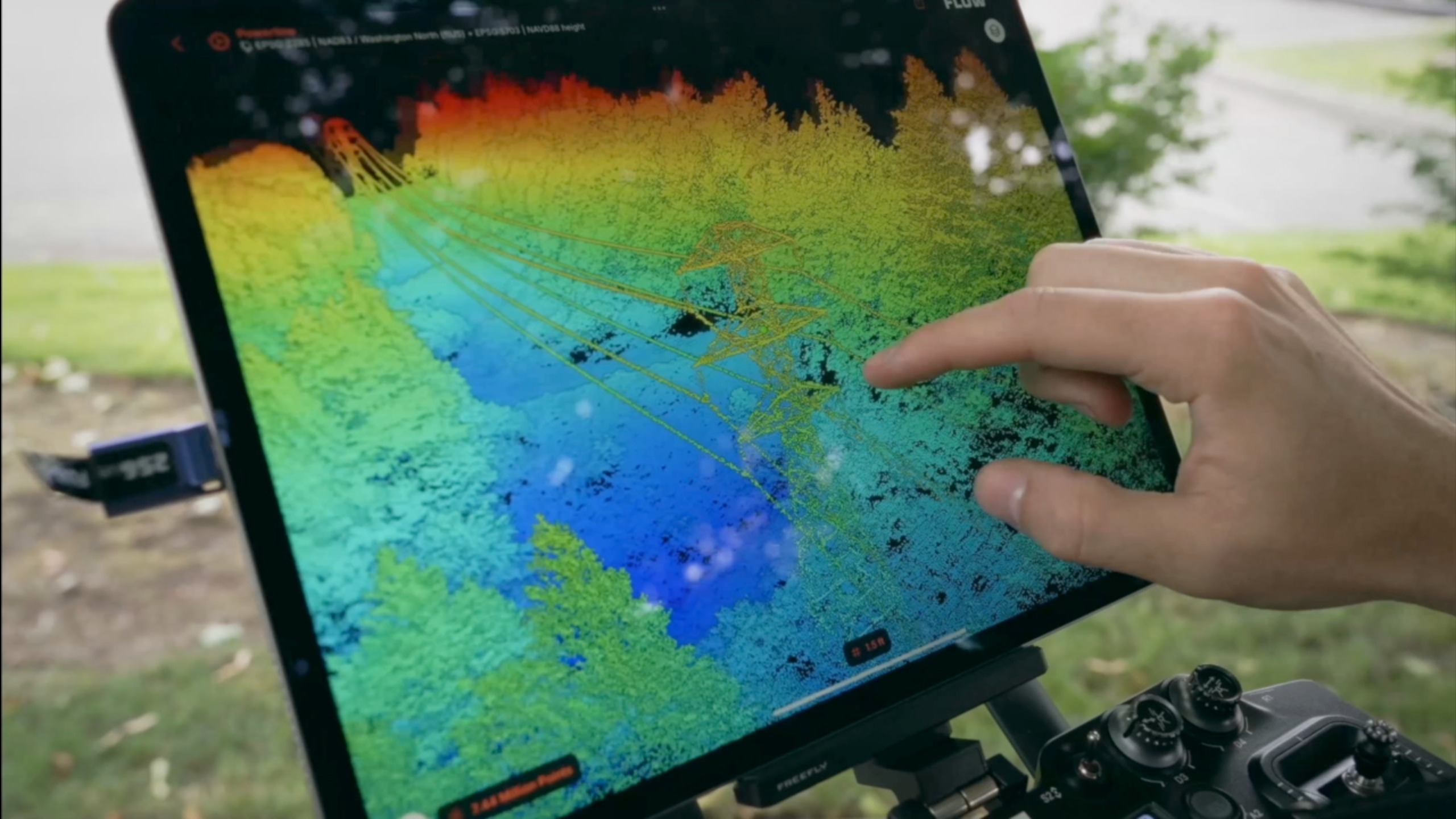 Freefly Flow tablet in the field displaying a colorized 2.44-million-point LiDAR point cloud of a transmission tower, conductors, and surrounding tree canopy for vegetation-encroachment review