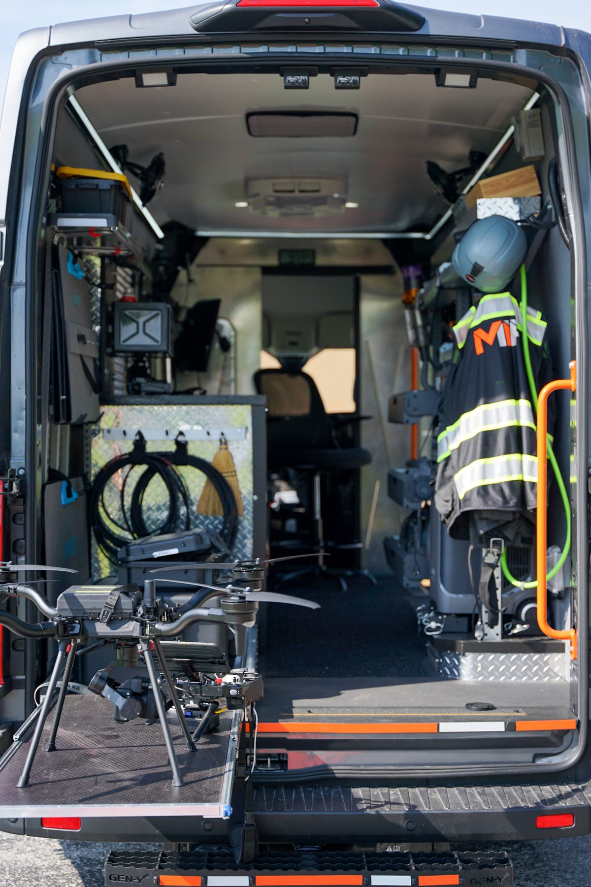 Open rear of the MIR Ford Transit command van at a solar site showing organized equipment, charging, the MIR-branded safety vest, and the drone on its deployment ramp