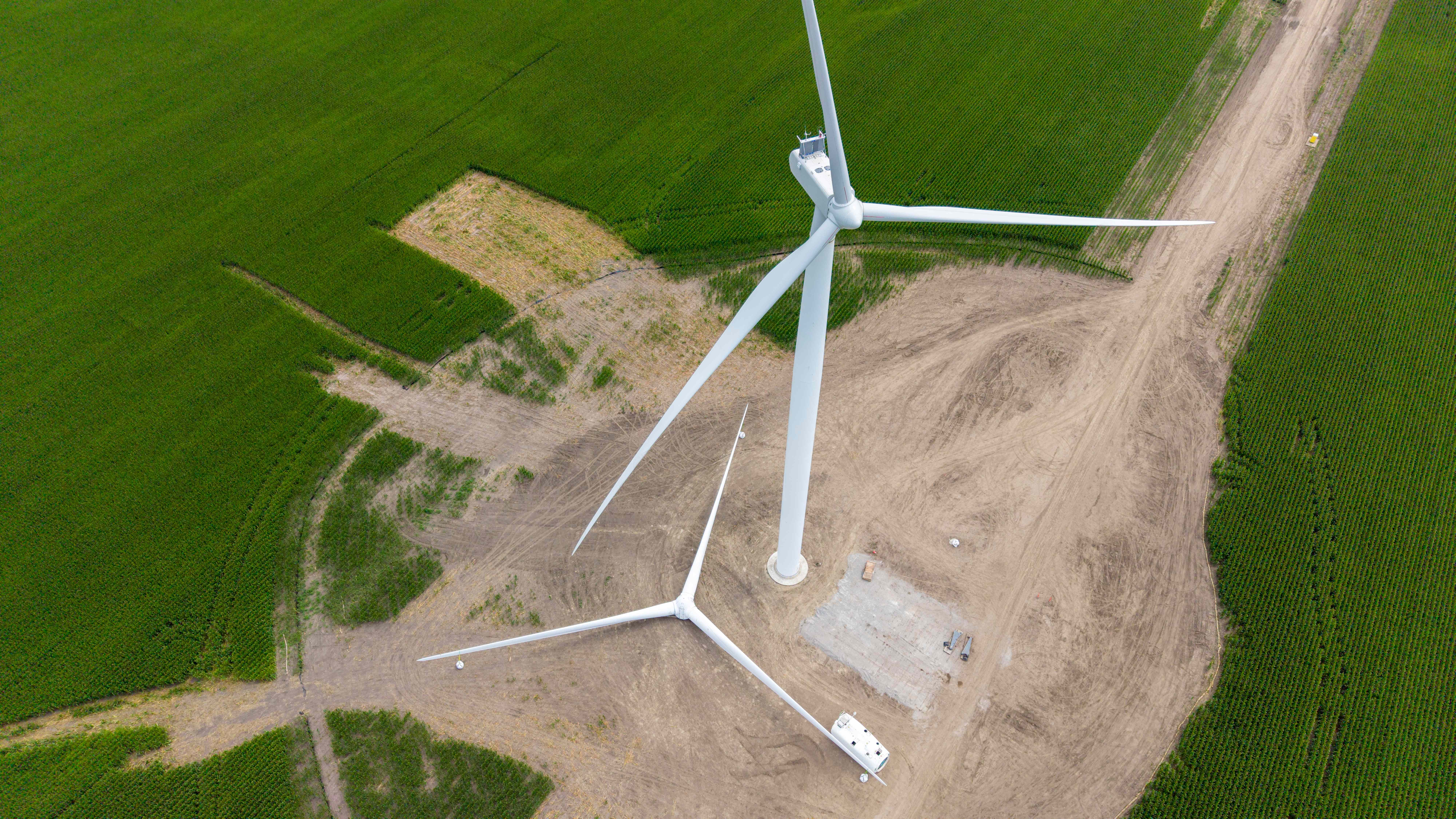Top-down aerial of a repower pad showing the turbine, foundation, crane paths, and access roads cut into surrounding crop fields, with a patch of disturbed and damaged crop measurable in the adjacent field