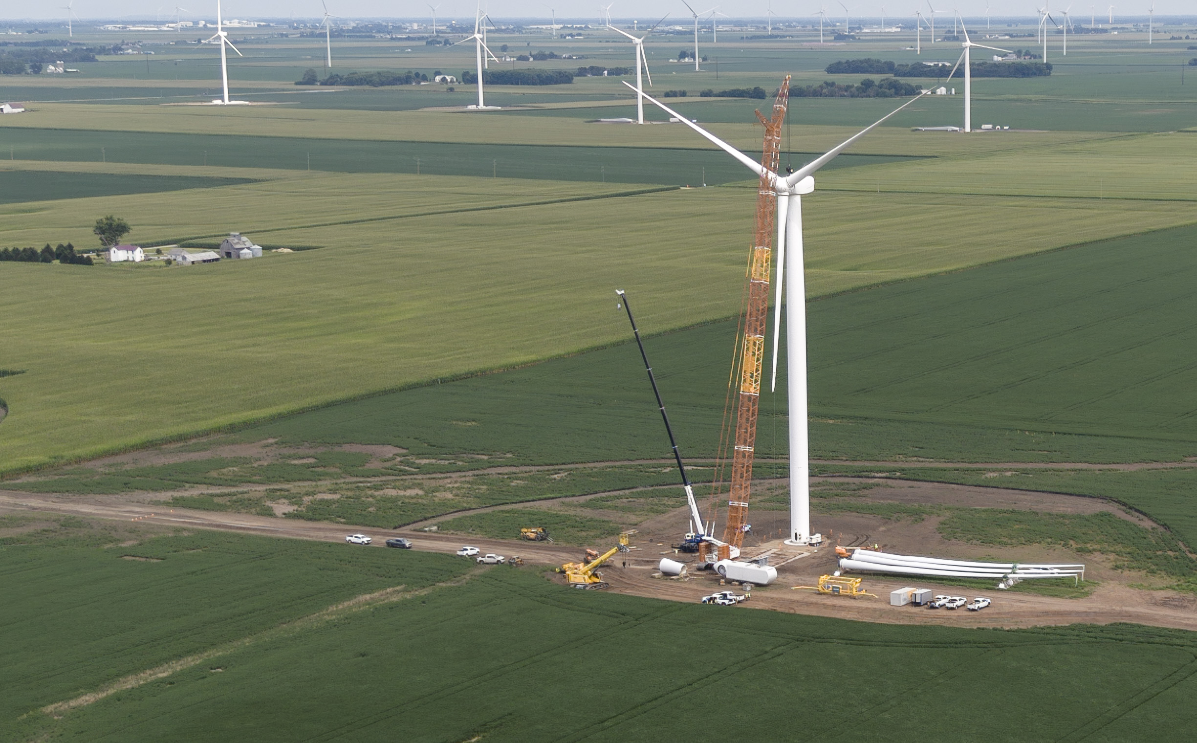 Aerial view of a wind turbine being erected during a repower — a crawler crane lifts the new rotor onto the tower while blades and components are staged on the graded pad, with existing turbines across the farmland behind