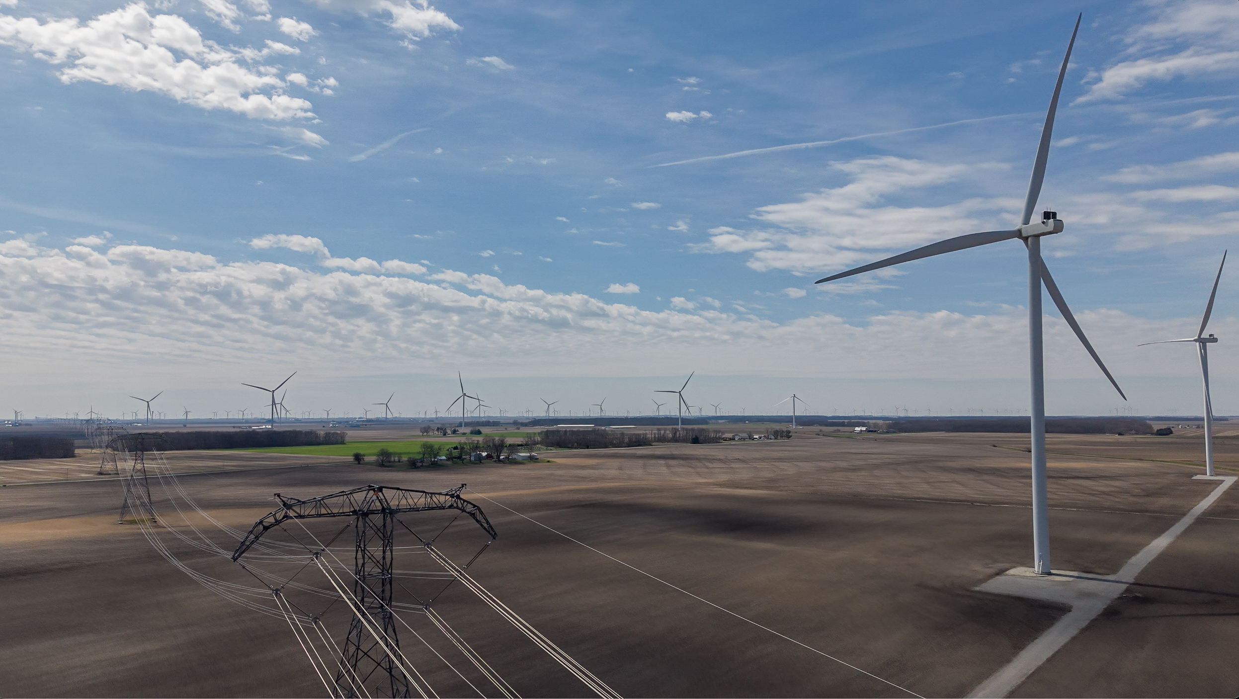 Aerial view of a utility-scale wind turbine rising over farmland beside a high-voltage transmission line, with a full wind farm receding to the horizon under a partly cloudy sky