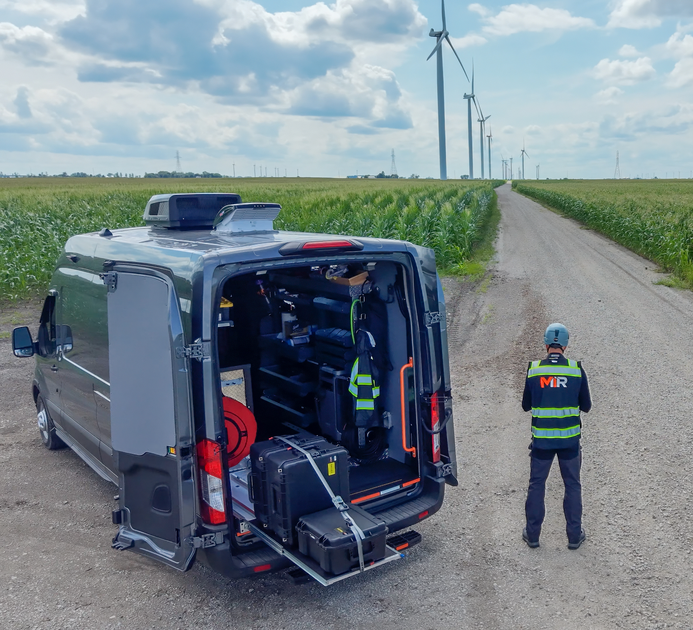 Andrew Hawes of MIR standing beside the mobile command van on a field road, drone equipment staged in the open rear doors, with wind turbines on the horizon